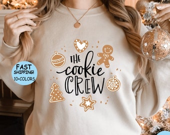 May include: A beige sweatshirt with a Christmas cookie-themed design. The text "The Cookie Crew" is printed in black with gingerbread cookies, ornaments, and snowflakes surrounding the text.