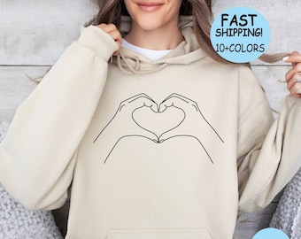 May include: A beige hoodie with a black outline of two hands forming a heart shape. The hoodie has a drawstring hood and a kangaroo pocket. The text "SAND" is printed on the bottom right corner of the hoodie.