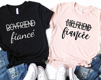 May include: Two t-shirts, one black and one pink, with the words "Boyfriend fiancé" and "Girlfriend fiancée" printed on them in white and black, respectively. The shirts are paired with blue jeans and white sneakers.