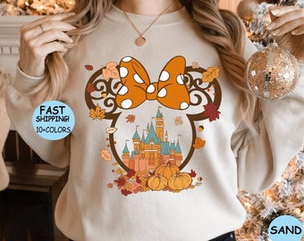 May include: A beige sweatshirt with a graphic of a cartoon mouse head with a bow and a castle. The bow is orange and white with polka dots. The castle is surrounded by fall leaves and pumpkins.