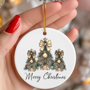 May include: A white ceramic Christmas ornament with three Christmas trees. The trees have gold bows and leopard print accents. The ornament has a gold string for hanging and the words "Merry Christmas" in black script.