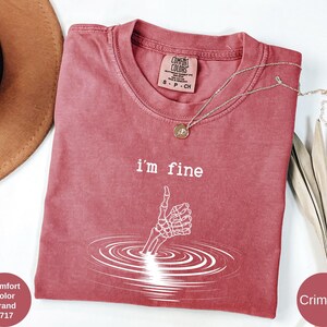 May include: Crimson-colored t-shirt with the text "I'm fine" and a white skeleton hand giving a thumbs up. The graphic is surrounded by concentric circles. A gold necklace is draped across the shirt.