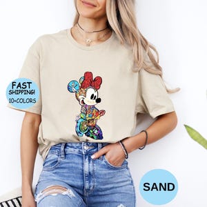 May include: A sand-colored t-shirt featuring a colorful graphic of Minnie Mouse. The graphic includes a red bow, and the text "FAST SHIPPING! 10+ COLORS" is visible on the left side. The word "SAND" is in a blue circle.