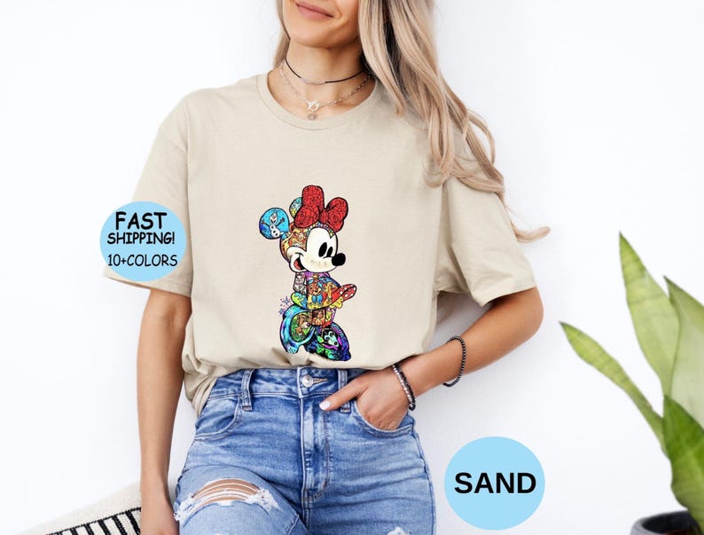 May include: A sand-colored t-shirt featuring a colorful graphic of Minnie Mouse. The graphic includes a red bow, and the text "FAST SHIPPING! 10+ COLORS" is visible on the left side. The word "SAND" is in a blue circle.