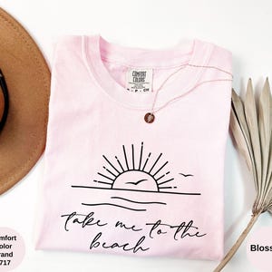 K&ouml;nnte beinhalten: Ein hellrosa Comfort Colors T-Shirt mit einer schwarzen Grafik einer Sonne &uuml;ber dem Meer und den Worten "Take me to the beach". Eine goldene Halskette mit Anh&auml;nger befindet sich auf dem Shirt.