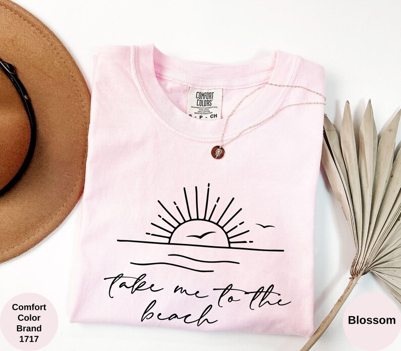 K&ouml;nnte beinhalten: Ein hellrosa Comfort Colors T-Shirt mit einer schwarzen Grafik einer Sonne &uuml;ber dem Meer und den Worten "Take me to the beach". Eine goldene Halskette mit Anh&auml;nger befindet sich auf dem Shirt.