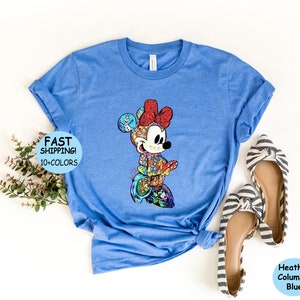 May include: Heather Columbia Blue t-shirt featuring a colorful Minnie Mouse design. The graphic is composed of various Disney characters and elements. The shirt has rolled-up sleeves and the text "FAST SHIPPING! 10+COLORS" is visible.