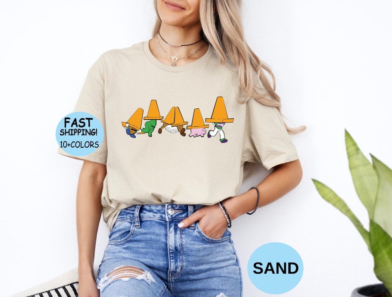 以下が含まれることがあります： サンドカラーのクルーネックTシャツ。頭にオレンジ色の三角コーンを乗せた5人のキャラクターが並んで歩くグラフィックがプリントされています。シャツには「FAST SHIPPING! 10+COLORS」と書かれた青い円形のバッジが含まれています。