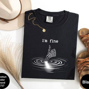 May include: Black t-shirt with the text "i'm fine" and a skeleton hand giving a thumbs up. The graphic is white. The shirt is next to a beige hat, gold necklace, and brown sandals.