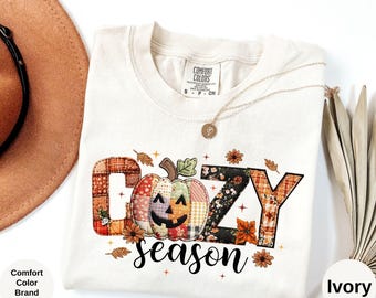Op de afbeelding: Ivoor Comfort Colors T-shirt met een "Cozy Season" ontwerp. Het ontwerp heeft een patchwork pompoen en letters, met herfstthema accenten. Het shirt heeft een ronde hals en korte mouwen.