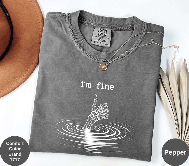 May include: Pepper-colored Comfort Colors brand t-shirt with the text "I'm fine" and a skeleton hand giving a thumbs up. The graphic is white and shows the hand emerging from water. A gold necklace is draped on the shirt.