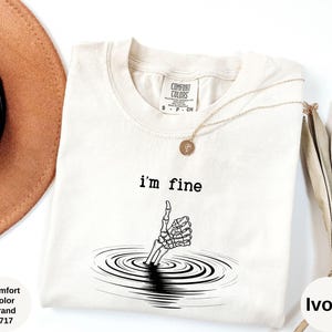 May include: Ivory-colored Comfort Colors brand t-shirt with the text "I'm fine" above a skeleton hand giving a thumbs up. The hand is emerging from a series of concentric circles. A gold necklace and a hat are visible.
