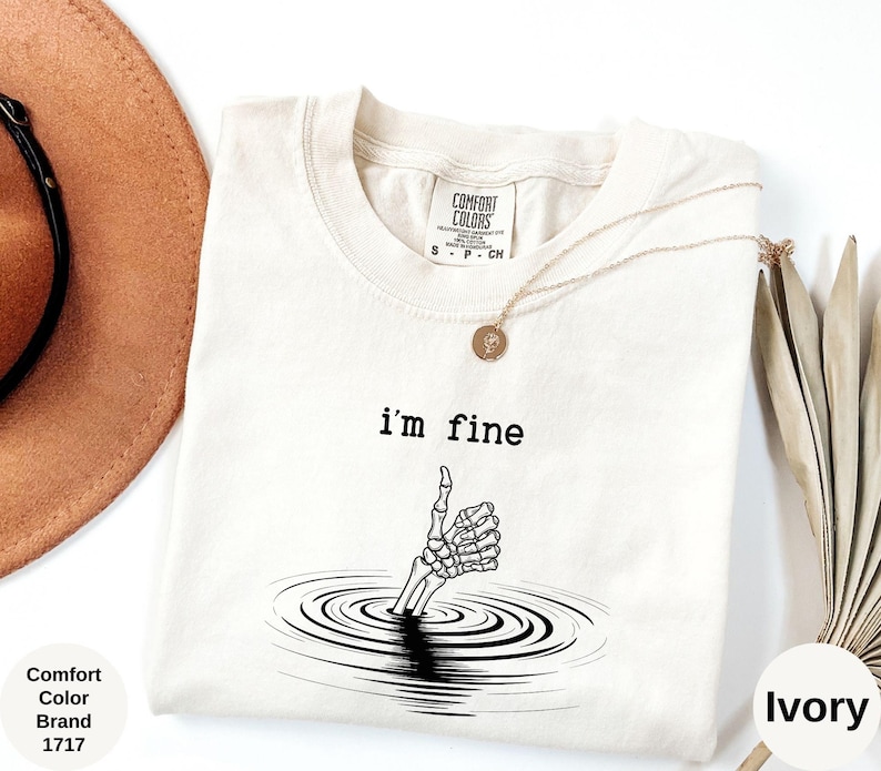 May include: Ivory-colored Comfort Colors brand t-shirt with the text "I'm fine" above a skeleton hand giving a thumbs up. The hand is emerging from a series of concentric circles. A gold necklace and a hat are visible.
