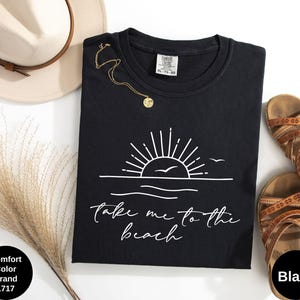K&ouml;nnte beinhalten: Schwarzes T-Shirt mit Rundhalsausschnitt und wei&szlig;er Grafik einer Sonne &uuml;ber dem Meer und den Worten "take me to the beach". Das Shirt liegt neben einem beigen Hut, einer goldenen Halskette und braunen Sandalen.