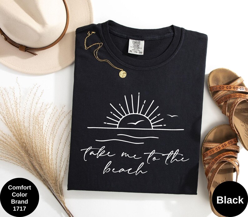 K&ouml;nnte beinhalten: Schwarzes T-Shirt mit Rundhalsausschnitt und wei&szlig;er Grafik einer Sonne &uuml;ber dem Meer und den Worten "take me to the beach". Das Shirt liegt neben einem beigen Hut, einer goldenen Halskette und braunen Sandalen.