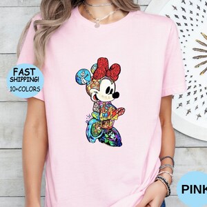 May include: A light pink t-shirt featuring a colorful Minnie Mouse design. The Minnie Mouse graphic is detailed with various patterns and colors. The shirt also has the text "FAST SHIPPING! 10+ COLORS" in a blue circle and the word "PINK" in a blue circle.