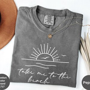 K&ouml;nnte beinhalten: Ein graues Comfort Colors T-Shirt mit einer wei&szlig;en Grafik einer Sonne &uuml;ber dem Meer und dem Text "Take me to the beach". Eine silberne Halskette mit Anh&auml;nger befindet sich auf dem T-Shirt. Das T-Shirt ist mit "Pepper" gekennzeichnet.