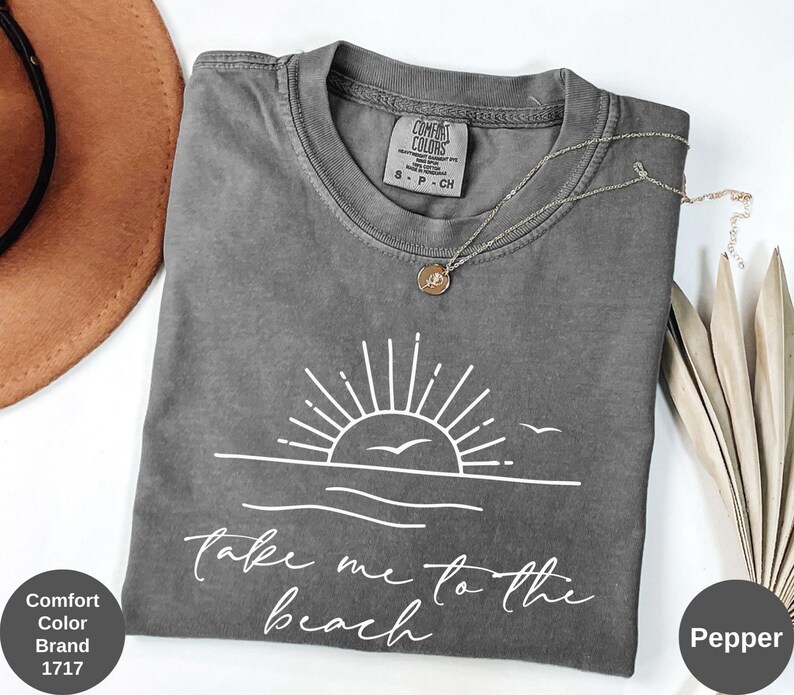 K&ouml;nnte beinhalten: Ein graues Comfort Colors T-Shirt mit einer wei&szlig;en Grafik einer Sonne &uuml;ber dem Meer und dem Text "Take me to the beach". Eine silberne Halskette mit Anh&auml;nger befindet sich auf dem T-Shirt. Das T-Shirt ist mit "Pepper" gekennzeichnet.