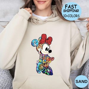 May include: A sand-colored hoodie featuring a colorful Minnie Mouse graphic. The Minnie Mouse design is composed of various characters and patterns. The hoodie has a front pocket and a drawstring hood. Text on the image says "FAST SHIPPING! 10+COLORS" and "SAND".