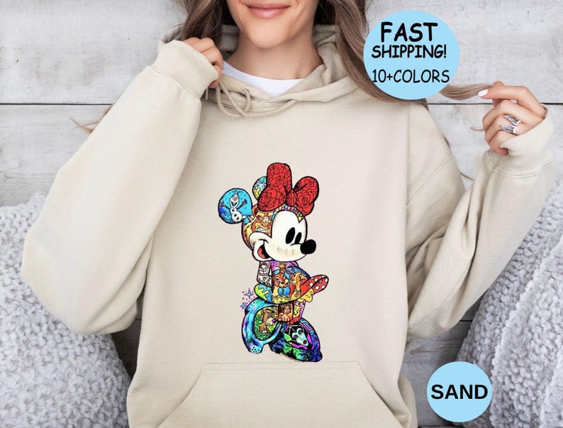 May include: A sand-colored hoodie featuring a colorful Minnie Mouse graphic. The Minnie Mouse design is composed of various characters and patterns. The hoodie has a front pocket and a drawstring hood. Text on the image says "FAST SHIPPING! 10+COLORS" and "SAND".