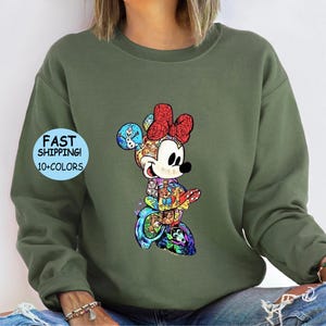 May include: Olive green sweatshirt featuring a colorful Minnie Mouse graphic. The Minnie Mouse design includes a red bow, and various character illustrations. The sweatshirt has the text "FAST SHIPPING! 10+ COLORS" on the left side and "MILITARY GREEN" on the right side.