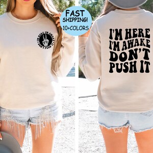 May include: A beige sweatshirt with the text "I'm here I'm awake Don't push it" printed in black on the back. The front of the sweatshirt has a small circle with a peace sign and the text "I'm here I'm awake Don't push it" printed in black.