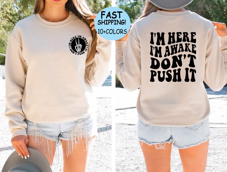 May include: A beige sweatshirt with the text "I'm here I'm awake Don't push it" printed in black on the back. The front of the sweatshirt has a small circle with a peace sign and the text "I'm here I'm awake Don't push it" printed in black.