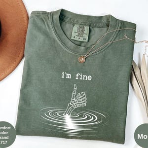 May include: Moss green Comfort Colors t-shirt with the text "I'm fine" and a white skeleton hand giving a thumbs up. A gold necklace with a pendant is draped across the shirt. The shirt is part of the Comfort Color Brand, style 1717.