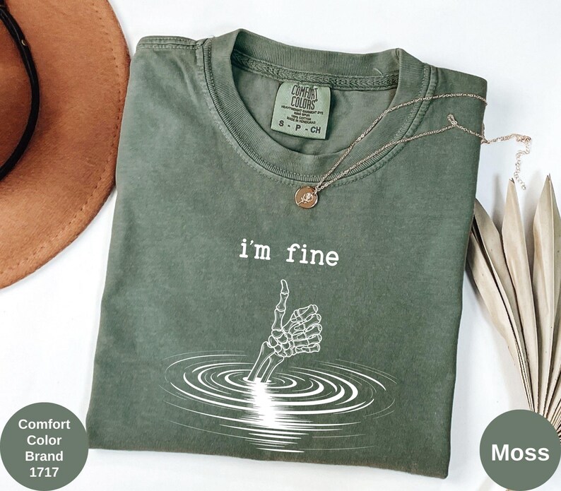 May include: Moss green Comfort Colors t-shirt with the text "I'm fine" and a white skeleton hand giving a thumbs up. A gold necklace with a pendant is draped across the shirt. The shirt is part of the Comfort Color Brand, style 1717.