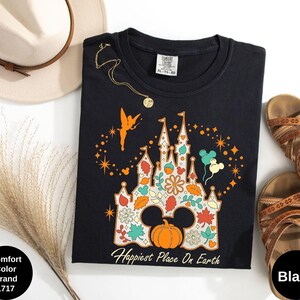 May include: Black crew neck t-shirt featuring a colorful castle design with a pumpkin, leaves, and Mickey Mouse ears. The text "Happiest Place On Earth" is printed below the castle. A gold necklace and sandals are next to the shirt.