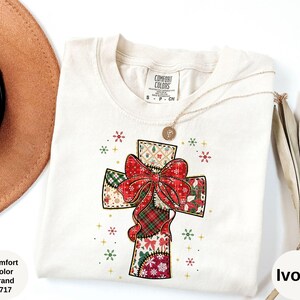 以下が含まれることがあります： クリスマス十字架のデザインが施された、アイボリー色のComfort ColorsブランドのTシャツ。十字架には大きな赤いリボンと、赤、緑、白のパッチワーク模様が施されています。Tシャツはクルーネックで半袖です。「Comfort Color Brand 1717」の文字が見えます。