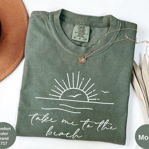 K&ouml;nnte beinhalten: Ein moosgr&uuml;nes Comfort Colors T-Shirt mit einer wei&szlig;en Grafik einer Sonne &uuml;ber dem Meer und den Worten "Take me to the beach". Eine kleine goldene Halskette liegt auf dem Shirt. Das Shirt ist Gr&ouml;&szlig;e S-P-CH.