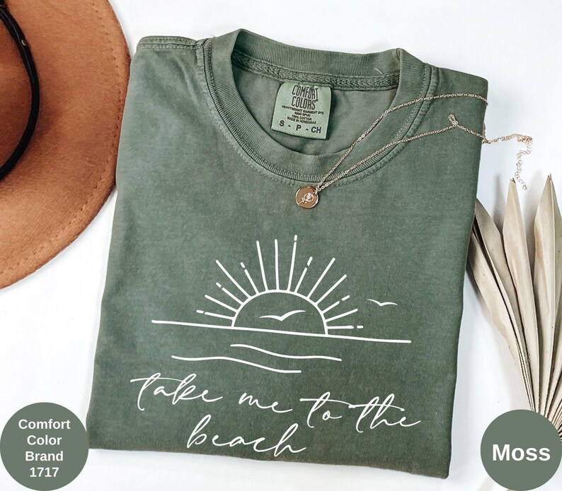 K&ouml;nnte beinhalten: Ein moosgr&uuml;nes Comfort Colors T-Shirt mit einer wei&szlig;en Grafik einer Sonne &uuml;ber dem Meer und den Worten "Take me to the beach". Eine kleine goldene Halskette liegt auf dem Shirt. Das Shirt ist Gr&ouml;&szlig;e S-P-CH.