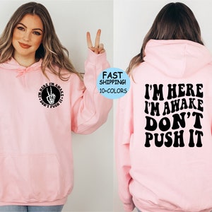 May include: Pink hoodie with a black graphic on the front that says "I'm here I'm awake don't push it" with a peace sign and skeleton hand. The back of the hoodie has the same text in a larger font.