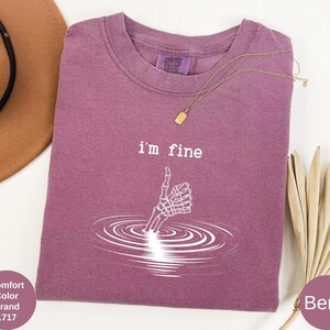 May include: Berry-colored t-shirt with the text "i'm fine" and a skeleton hand giving a thumbs up. The graphic is white and depicts the hand emerging from water. The shirt is a Comfort Colors brand.