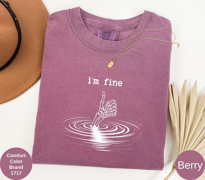 May include: Berry-colored t-shirt with the text "i'm fine" and a skeleton hand giving a thumbs up. The graphic is white and depicts the hand emerging from water. The shirt is a Comfort Colors brand.