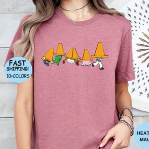 以下が含まれることがあります： ヘザーモーブ色の半袖Tシャツ。頭にオレンジ色の三角コーンを被った5人のキャラクターのグラフィックプリントが施されています。キャラクターは一列に並んでおり、それぞれがコーンによって部分的に隠れているデザインです。