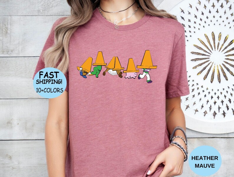 以下が含まれることがあります： ヘザーモーブ色の半袖Tシャツ。頭にオレンジ色の三角コーンを被った5人のキャラクターのグラフィックプリントが施されています。キャラクターは一列に並んでおり、それぞれがコーンによって部分的に隠れているデザインです。