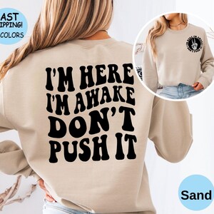 May include: Sand colored sweatshirt with the text "I'm here I'm awake Don't push it" printed in black.