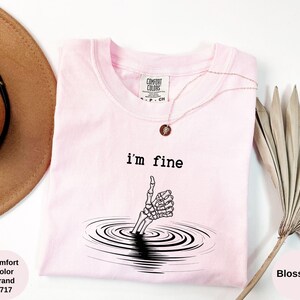 May include: A light pink Comfort Colors t-shirt with the text "i'm fine" above a skeleton hand emerging from water. A gold necklace with a pendant is on the shirt. A brown hat and dried plant are in the background.