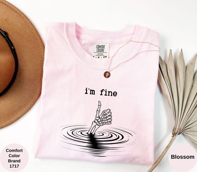 May include: A light pink Comfort Colors t-shirt with the text "i'm fine" above a skeleton hand emerging from water. A gold necklace with a pendant is on the shirt. A brown hat and dried plant are in the background.
