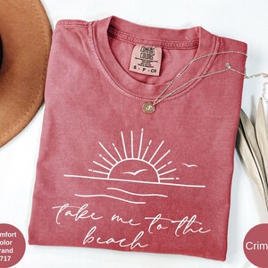 K&ouml;nnte beinhalten: Karmesinrotes T-Shirt mit wei&szlig;er Grafik von Sonne, Wellen und V&ouml;geln sowie dem Text "take me to the beach". Eine goldene Halskette liegt auf dem Shirt. Das Shirt ist von der Marke Comfort Colors.