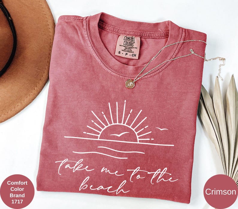 K&ouml;nnte beinhalten: Karmesinrotes T-Shirt mit wei&szlig;er Grafik von Sonne, Wellen und V&ouml;geln sowie dem Text "take me to the beach". Eine goldene Halskette liegt auf dem Shirt. Das Shirt ist von der Marke Comfort Colors.
