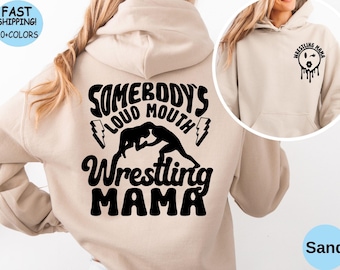 May include: A beige hoodie with a black graphic that says "Somebody's Loud Mouth Wrestling Mama" with a silhouette of two women wrestling. The hoodie has a pocket on the front with a black graphic of a cartoon face with a kissy face and the words "Wrestling Mama".