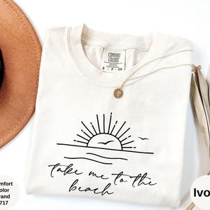 K&ouml;nnte beinhalten: Elfenbeinfarbenes T-Shirt mit einer schwarzen Grafik einer Sonne &uuml;ber dem Meer und dem Text "Take me to the beach". Das Shirt ist von der Marke Comfort Colors, Stil 1717. Eine goldene Halskette und ein brauner Hut sind ebenfalls sichtbar.