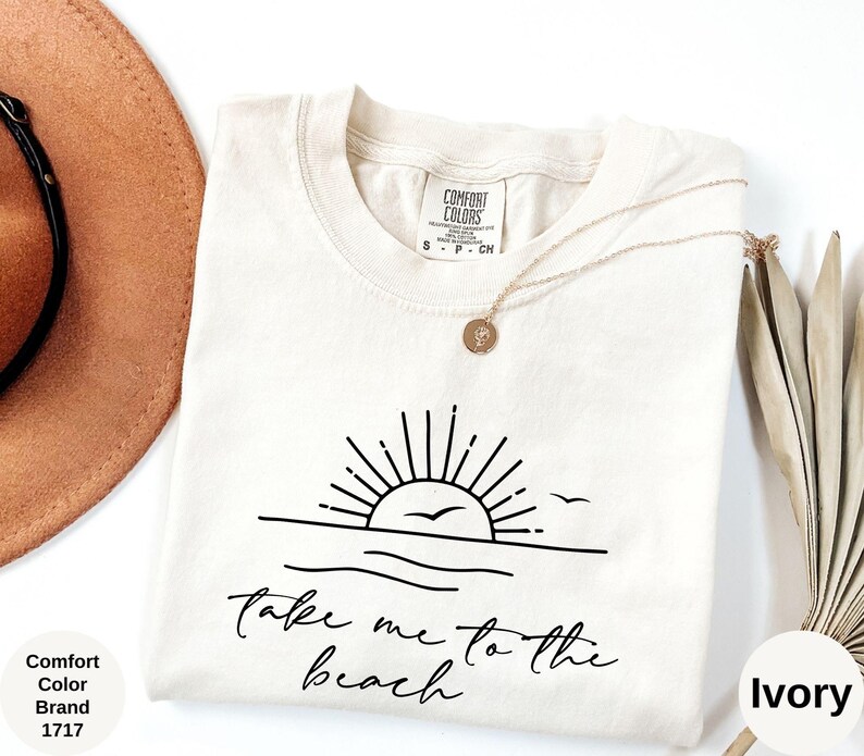 K&ouml;nnte beinhalten: Elfenbeinfarbenes T-Shirt mit einer schwarzen Grafik einer Sonne &uuml;ber dem Meer und dem Text "Take me to the beach". Das Shirt ist von der Marke Comfort Colors, Stil 1717. Eine goldene Halskette und ein brauner Hut sind ebenfalls sichtbar.