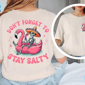 Puede incluir: Una camiseta blanca con el texto "Don't forget to stay salty" y un gráfico de un esqueleto con un sombrero y montando un flamenco rosa con un cóctel.