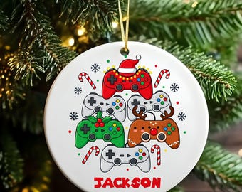 May include: A white, round Christmas ornament featuring a collection of game controllers in various colors. The controllers are decorated with festive elements like a Santa hat, holly, and reindeer antlers. The name "JACKSON" is printed in red.