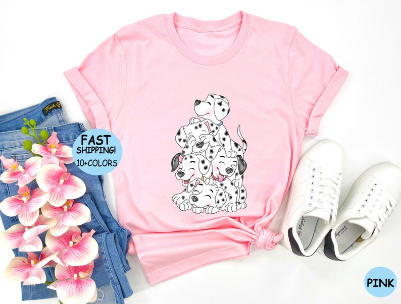 Puede incluir: Camiseta rosa con un gr&aacute;fico de cachorros d&aacute;lmatas apilados. Los cachorros son blancos y negros con caras sonrientes. La camiseta tiene las mangas enrolladas y el texto "FAST SHIPPING! 10+ COLORS".
