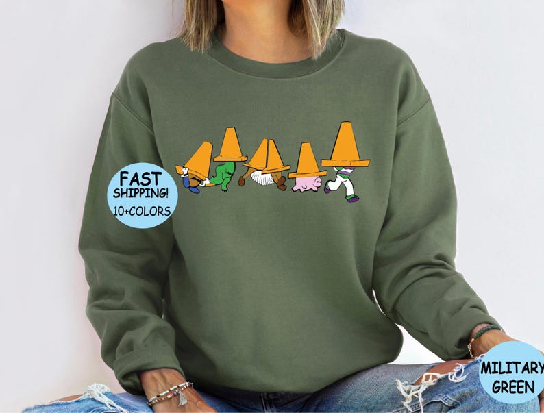 May include: A green sweatshirt with a graphic of five people wearing traffic cones on their heads. The graphic is in black, white, and orange. The text "FAST SHIPPING! 10+ COLORS" is on a blue circle in the lower left corner of the image.